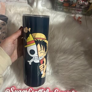 May include: A black and white anime character with a straw hat is printed on a black tumbler. The tumbler is held in a hand. The image also includes the text "Sugar Crafts by Graciela" and social media icons for Facebook and Instagram.