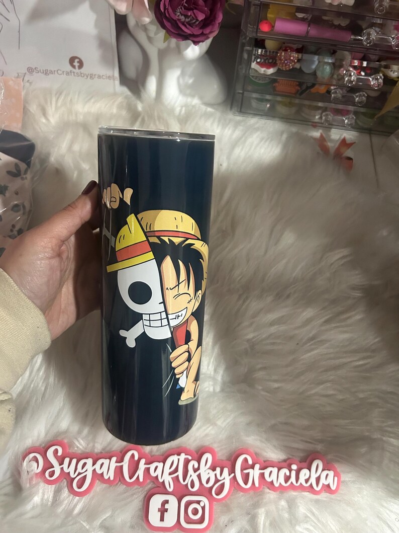 May include: A black and white anime character with a straw hat is printed on a black tumbler. The tumbler is held in a hand. The image also includes the text "Sugar Crafts by Graciela" and social media icons for Facebook and Instagram.