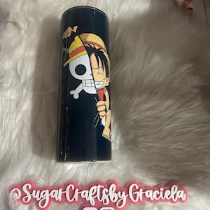 May include: A black and white anime character with a straw hat is printed on a black tumbler. The character is holding a skull and crossbones. The tumbler is on a white fuzzy surface.