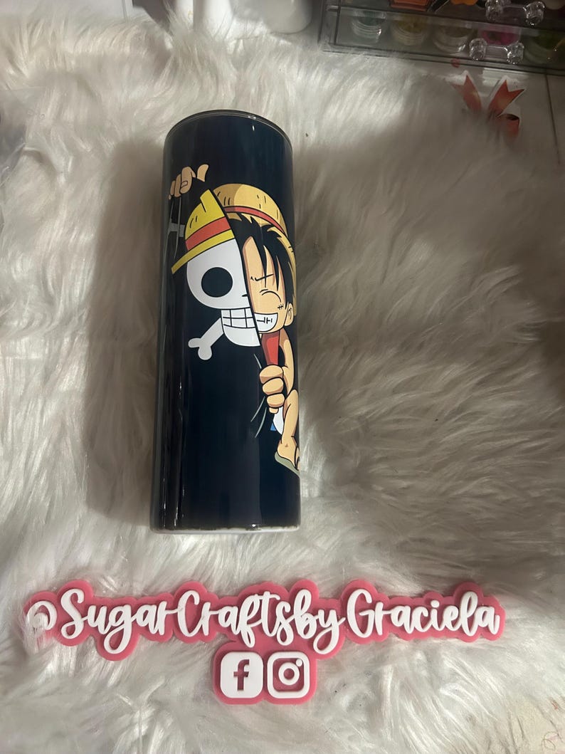 May include: A black and white anime character with a straw hat is printed on a black tumbler. The character is holding a skull and crossbones. The tumbler is on a white fuzzy surface.