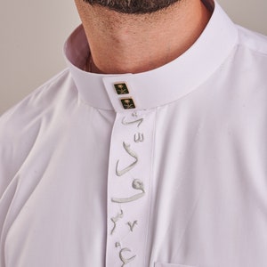 Puede incluir: Camisa blanca de manga larga con cuello alto y una fila vertical de bordados plateados. La camisa tiene dos botones pequeños de oro con emblemas verdes y dorados en el cuello.