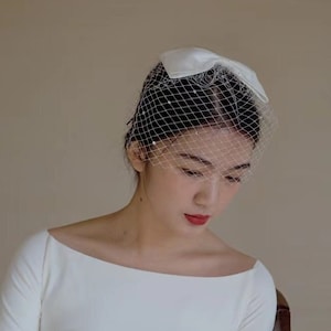 May include: A woman wearing a white dress with long sleeves and a white veil with pearl accents. She is sitting on a wooden chair and looking down.