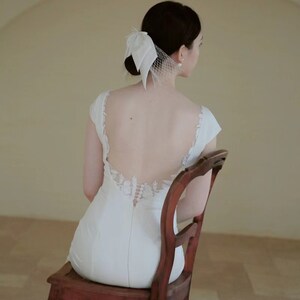 May include: A woman wearing a white wedding dress with a lace back detail sits on a wooden chair. She is wearing a white veil with a bow on the back of her head.