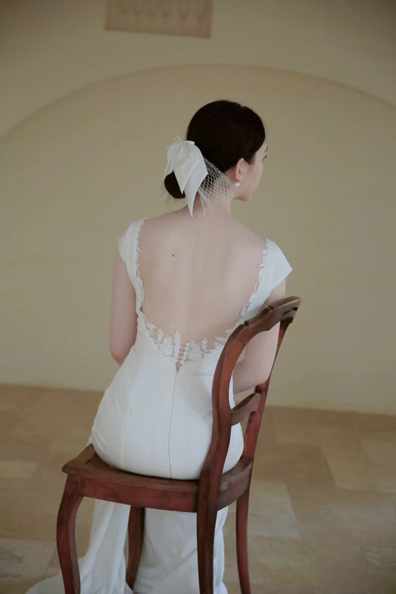 May include: A woman wearing a white wedding dress with a lace back detail sits on a wooden chair. She is wearing a white veil with a bow on the back of her head.
