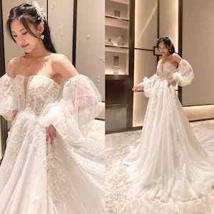 Op de afbeelding: Een witte off-shoulder bruidsjurk met een strakke buste en een zwierige rok. De jurk heeft delicate kantdetails en pofmouwen.
