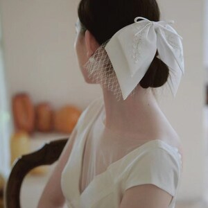 May include: A white bridal hair accessory with a large bow and a veil. The bow is made of satin and has a delicate lace design. The veil is made of tulle and is attached to the bow.