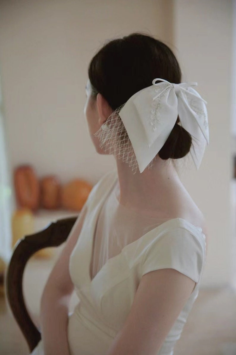 May include: A white bridal hair accessory with a large bow and a veil. The bow is made of satin and has a delicate lace design. The veil is made of tulle and is attached to the bow.