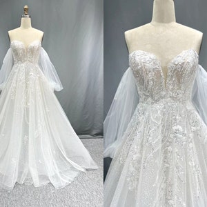 Op de afbeelding: Een witte off-shoulder bruidsjurk met een sweetheart-halslijn en een aansluitend lijfje. De jurk heeft een volle rok met een tule-overlay en delicate bloemenborduursels. De jurk heeft lange, pofmouwen van een transparante stof.
