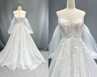 May include: A white off-the-shoulder wedding dress with a sweetheart neckline and a fitted bodice. The dress has a full skirt with a tulle overlay and delicate floral embroidery. The dress features long, puffy sleeves with a sheer fabric.