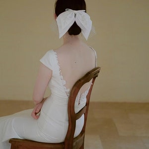 May include: A woman wearing a white wedding dress with a low back and a white bow in her hair. She is sitting on a wooden chair.