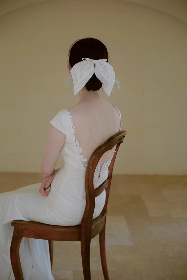 May include: A woman wearing a white wedding dress with a low back and a white bow in her hair. She is sitting on a wooden chair.
