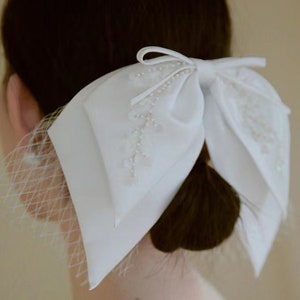 May include: A white satin bow with pearl embellishments and a delicate tulle veil. The bow is tied in a large, elegant knot and is perfect for adding a touch of sophistication to any hairstyle.