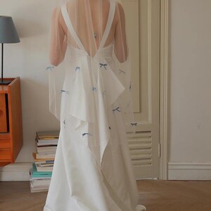Puede incluir: Un vestido de novia blanco con la espalda baja y una falda fluida. El vestido está adornado con pequeños lazos azules. Un velo transparente, también decorado con lazos azules, cubre los hombros. El vestido es elegante y adecuado para una boda.
