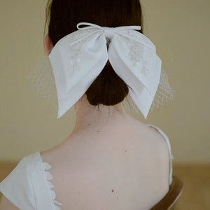 May include: A white satin bow with pearl embellishments, worn in the hair as a bridal accessory.