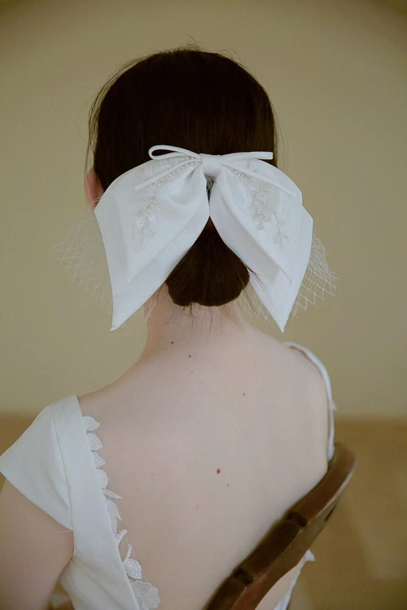 May include: A white satin bow with pearl embellishments, worn in the hair as a bridal accessory.