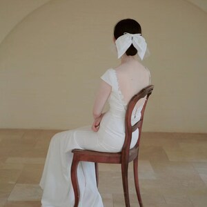 May include: A woman in a white wedding dress sits on a wooden chair with her back to the camera. She is wearing a white bow in her hair.