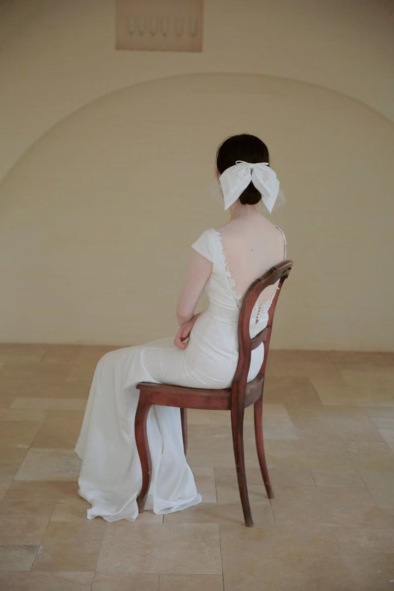 May include: A woman in a white wedding dress sits on a wooden chair with her back to the camera. She is wearing a white bow in her hair.