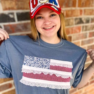 Op de afbeelding: Een blauw t-shirt met een rechthoekige vlag-geïnspireerde applicatie van blauwe bloemenstof, rood-witte vichy-stof en witte broderie anglaise. Een wit en rood trucker-petje met de letters USA in rood, wit en blauw.