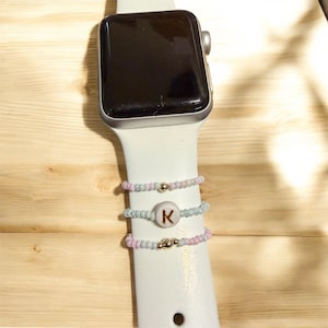 Puede incluir: Un reloj inteligente plateado con una correa blanca está adornado con tres anillos de cuentas. Los anillos presentan cuentas rosas y azules pastel, con una cuenta dorada. Un anillo tiene la letra "K".
