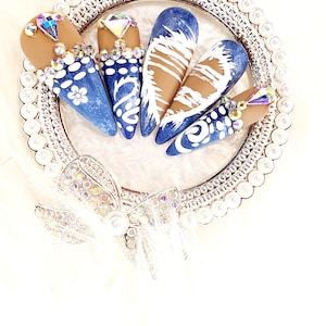 May include: Five blue and white nail tips with rhinestones on a white background. The nail tips are arranged in a circle on a silver tray with a pearl border.