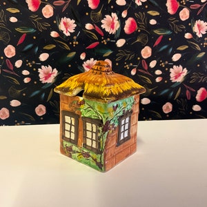 Op de afbeelding: Een keramische suikerpot in de vorm van een huisje met een bruin rieten dak, groene klimop en twee ramen. De pot is geschilderd in tinten bruin, groen en geel.