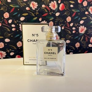 Può includere: Un flacone trasparente di Chanel No. 5 Eau de Parfum con forma rettangolare e tappo trasparente sfaccettato. Il flacone è accanto a una scatola bianca con testo nero che recita "N°5 CHANEL PARIS". Il flacone contiene 50 ml.