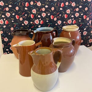 May include: A selection of ceramic jugs in shades of brown and beige. Some jugs feature a two-tone design, with a lighter base and darker top. The jugs are displayed on a white surface.