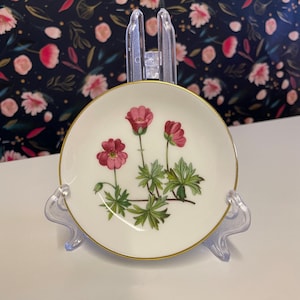 May include: A small white porcelain saucer with a gold rim features a floral design of three pink flowers with green leaves.