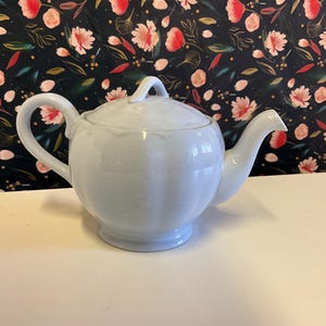 Może przedstawiać: Jasnoniebieski ceramiczny czajnik z pokrywką i zaokrąglonym korpusem. Czajnik ma prosty design z lekko uniesioną krawędzią wokół pokrywki.