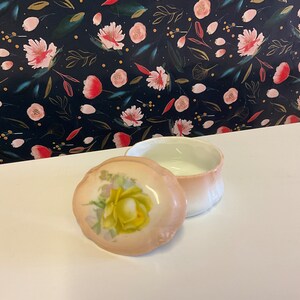 Może przedstawiać: Małe, otwarte, porcelanowe pudełko na drobiazgi z pokrywką. Pokrywka przedstawia malunek żółtej róży na brzoskwiniowym tle. Wnętrze pudełka jest białe i ma brzoskwiniową obwódkę. Tło to ciemny wzór kwiatowy.