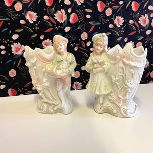 May include: A pair of antique porcelain vases. Each vase has a sculpted figure, one a man with a basket, the other a woman with flowers. The vases are white with pink and green accents, set against a floral patterned background.