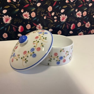 Op de afbeelding: Een witte porseleinen sieradendoos met een bloemenontwerp in tinten roze, blauw en groen. Het deksel heeft een blauwe knop en een witte rand.