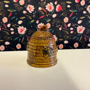 Könnte beinhalten: Ein brauner Keramik-Honigtopf in Form eines Bienenstocks mit Bienen an den Seiten.