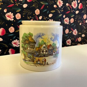 Può includere: Vaso cilindrico in ceramica color crema. Il vaso presenta una dettagliata illustrazione di una scena di villaggio europeo con edifici, alberi e figure. Il testo "Down Somerset Way" è stampato sul vaso.