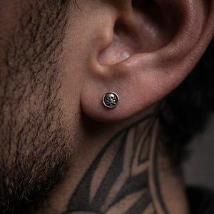 Puede incluir: Un pequeño pendiente de botón redondo plateado con un diseño de calavera y huesos cruzados. La imagen destaca el diseño del pendiente, adecuado para quienes buscan joyería.