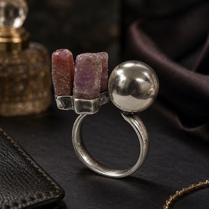May include: A silver ring featuring three rough-cut, reddish-purple gemstones and a polished silver sphere. The ring's band is a simple, elegant design, showcasing the unique texture and color of the stones.