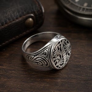 Könnte beinhalten: Ein silberner Ring mit ovaler Oberfläche und aufwendigem floralem Design. Das Band des Rings ist mit wirbelnden Mustern verziert, und das Metall hat eine polierte Oberfläche. Der Ring wird auf einer dunklen Holzoberfläche gezeigt.