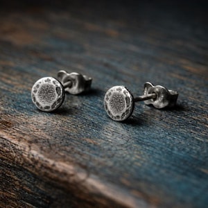 Può includere: Due orecchini a bottone argentati con un design circolare strutturato. Gli orecchini sono appoggiati su una superficie di legno blu e marrone. Gli orecchini hanno un perno e una chiusura a farfalla.