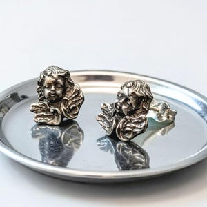 May include: A pair of silver-toned stud earrings, each featuring a detailed cherub face with closed eyes and delicate wings. The earrings are displayed on a small, round, silver tray, reflecting the light. The cherub design gives the earrings a vintage or antique aesthetic.