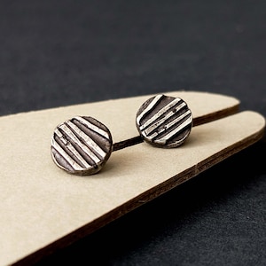 Op de afbeelding: Een paar kleine, ronde oorstekers. Elke oorbel is zilverkleurig met een textuur en diagonale lijnen. De oorbellen worden gepresenteerd op een lichtbruin oppervlak tegen een donkere achtergrond.