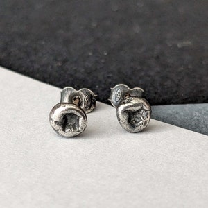 Peut inclure: Une paire de petites boucles d'oreilles rondes en argent. Chaque boucle d'oreille a un aspect texturé et vieilli avec une indentation carrée au centre. Les tiges des boucles d'oreilles sont visibles, avec un poinçon "925".