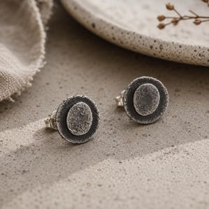 May include: A pair of silver stud earrings. Each earring features a textured, circular design with a raised, smaller circle in the center. The earrings have a dark, oxidized finish, giving them a vintage or antique appearance. The earrings are displayed on a neutral surface.
