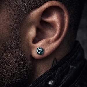 May include: Close-up of a teal-colored, textured ear stud. The stud is round with a slightly irregular surface, giving it a unique, handcrafted appearance. The earring is set in an ear, with a dark background.