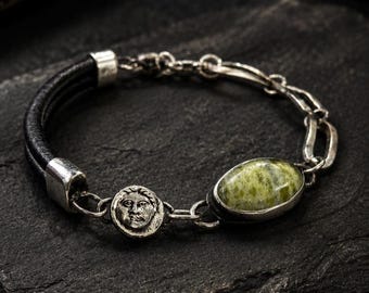 Puede incluir: Una pulsera de plata con una banda de cuero negro y una piedra ovalada verde. La pulsera presenta un pequeño medallón de plata con una cara y un diseño de eslabones de cadena. La pulsera está sobre una superficie negra.