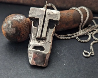 May include: A silver pendant necklace featuring a face-like design with a textured surface. The pendant is hanging from a silver chain.