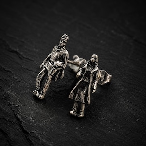 May include: A pair of silver-toned stud earrings. One earring depicts a man in a shirt and pants, the other a woman in a long coat. The earrings are set against a dark, textured background.