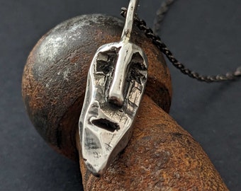 May include: A silver pendant necklace featuring a textured, abstract face design. The pendant is hanging from a black chain.
