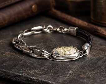 May include: Silver bracelet with a black leather band and a central oval stone. The bracelet features a chain link design on one side and a decorative stone set in a silver bezel. The stone has a cream and blue color.