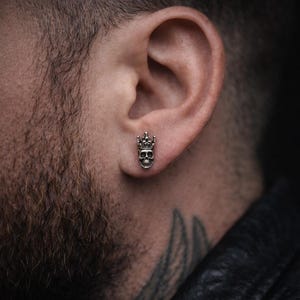 May include: A silver skull stud earring with a crown. The earring is worn on the ear of a person with a beard and a tattoo on their neck. The skull has a detailed design.