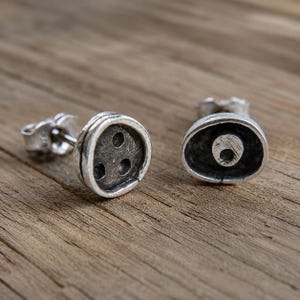 Puede incluir: Dos pendientes de plata. Uno tiene tres agujeros pequeños, el otro un círculo dentro de una forma ovalada. Los pendientes están sobre una superficie de madera.
